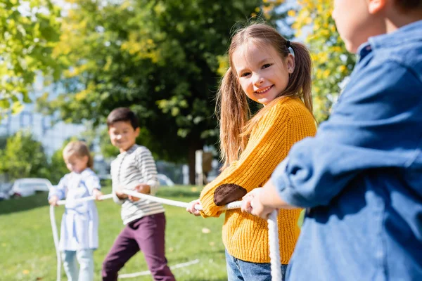 Ragazza sorridente guardando la fotocamera durante il tiro alla fune con gli amici su sfondo sfocato nel parco — Foto stock