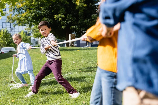 Asiático chico jugando tirón de la guerra con amigos en borrosa primer plano en hierba en parque - foto de stock