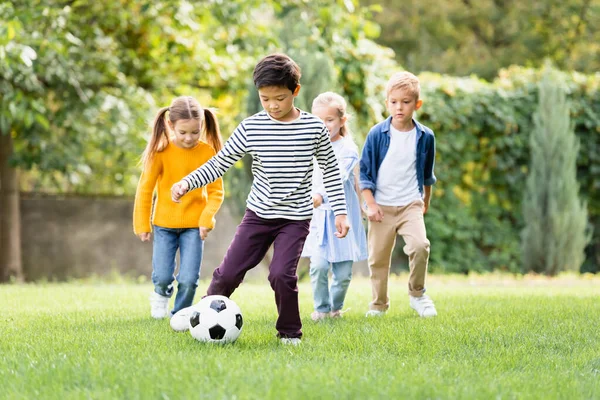 Asiatico ragazzo giocare calcio vicino amici su sfocato sfondo in parco — Foto stock