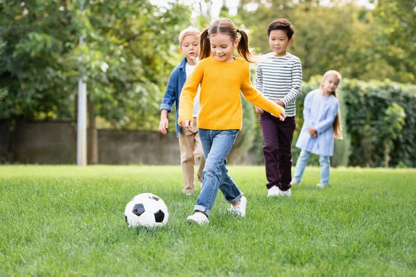 Lächelndes Mädchen spielt Fußball in der Nähe multiethnischer Freunde auf verschwommenem Hintergrund im Park — Stockfoto