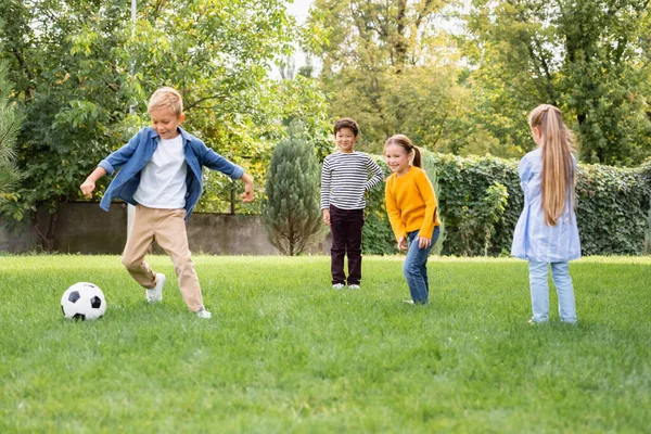 Positivos niños multiétnicos de pie cerca de un amigo jugando al fútbol en el parque - foto de stock