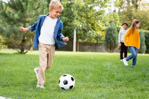 Sonriente niño jugando fútbol cerca de amigos en fondo borroso en el parque - foto de stock