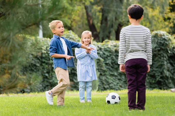 Excited boy pointing with fingers near football and friends in park — Stock Photo