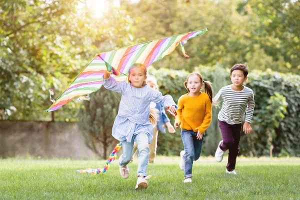 Chica sosteniendo volando cometa mientras corre cerca de amigos multiétnicos en el parque - foto de stock