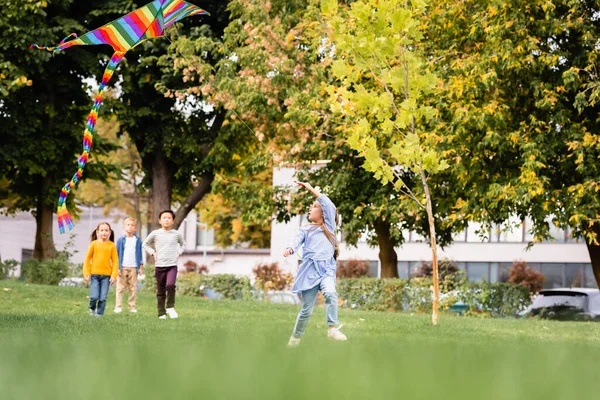 Girl playing with flying kite while multiethnic friends walking on grassy lawn on blurred background — Stock Photo