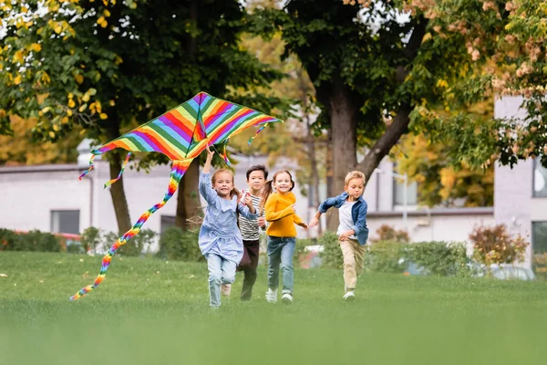Sonriendo niños multiétnicos corriendo por el césped mientras juegan con cometa voladora en el parque - foto de stock