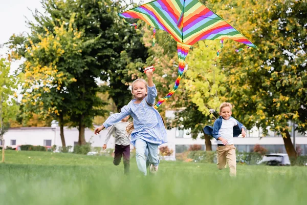 Sonriente chica sosteniendo volando cometa mientras jugando con los niños en fondo borroso en el parque - foto de stock