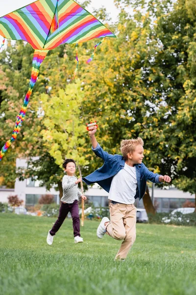 Smiling boy holding flying kite while running near asian friend in park — Stock Photo