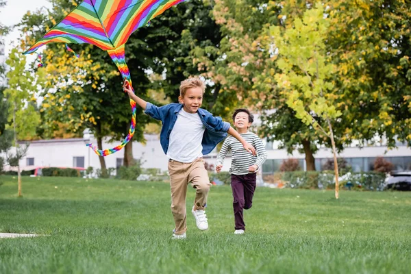 Cheerful boy running with flying kite while playing with asian friend in park — Stock Photo