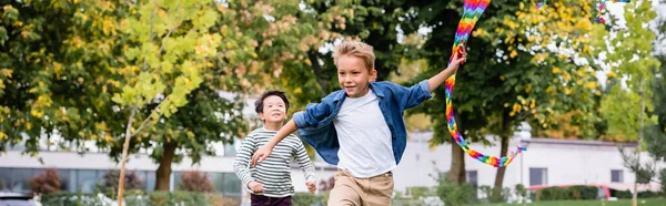 Sonriente niño sosteniendo colorido volando cometa mientras corriendo cerca asiático amigo en parque, bandera - foto de stock