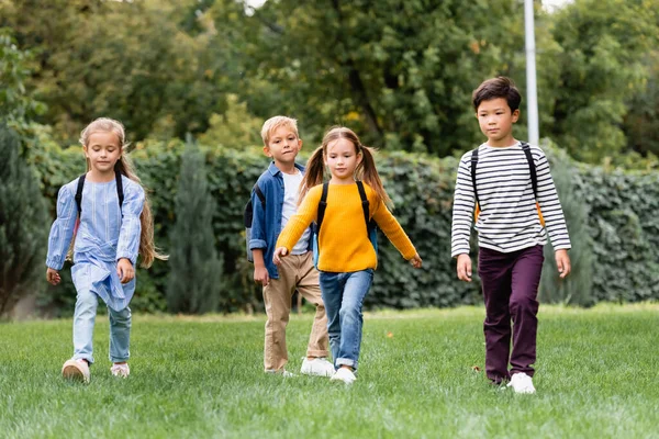 Scolari multiculturali che camminano con zaini sul prato nel parco — Foto stock