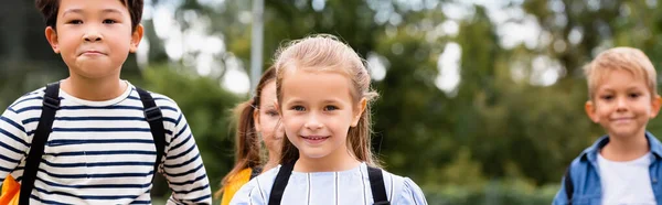 Fille souriante avec sac à dos regardant la caméra près d'amis multiethniques sur fond flou, bannière — Photo de stock