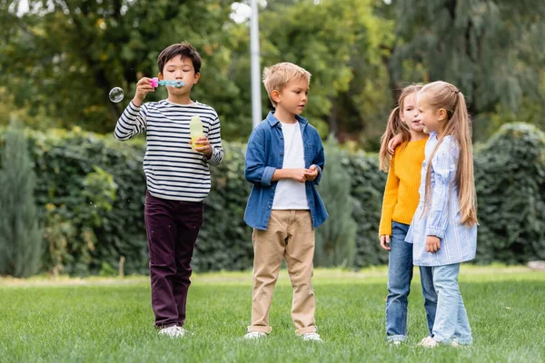 Asiático chico soplando jabón burbujas mientras de pie cerca sonriendo amigos en hierba césped - foto de stock