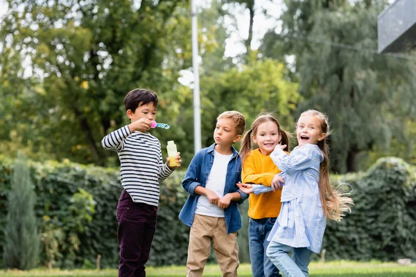 Bambini allegri multietnici con bolle di sapone in piedi nel parco — Foto stock