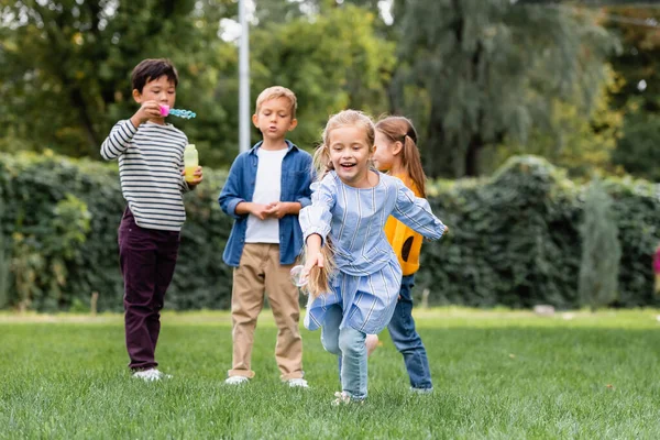 Chica sonriente corriendo en el césped cerca de amigos multiétnicos con burbujas de jabón sobre fondo borroso - foto de stock