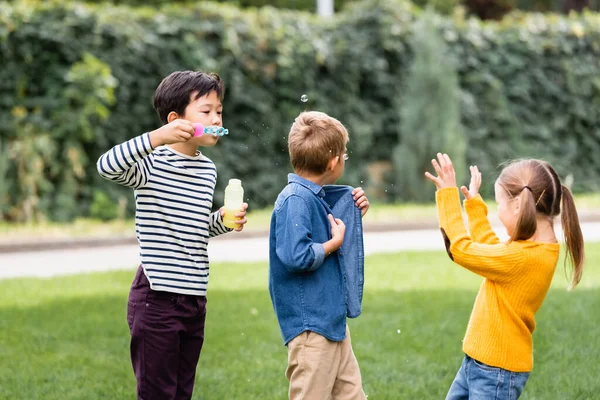 Asiatico scolaro soffiando sapone bolle vicino amici in parco — Foto stock