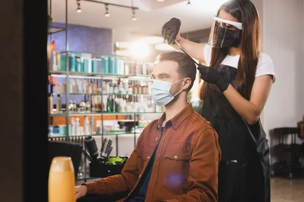 Barbiere in lattice guanti e viso scudo taglio capelli del cliente in maschera medica, sfocato primo piano — Foto stock