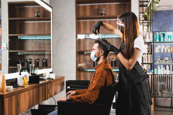 Vista lateral del peluquero en protector facial y guantes de látex cortando el pelo del cliente en máscara médica - foto de stock