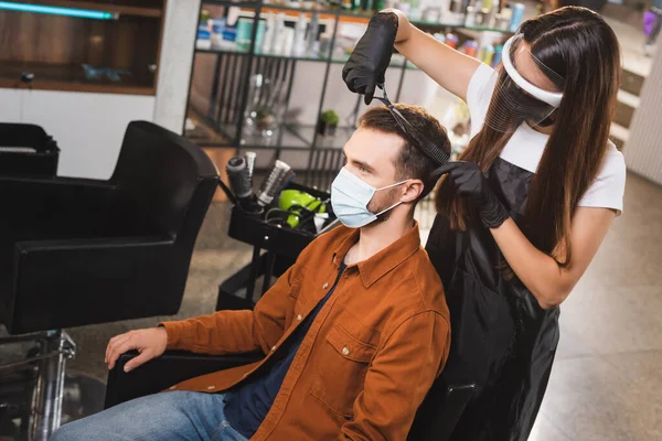 Barber in face shield and latex gloves cutting hair of client wearing medical mask — Stock Photo