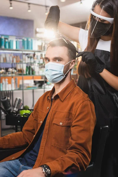 Мужчина в медицинской маске смотрит в камеру, в то время как парикмахер в защитном оборудовании стрижет волосы — стоковое фото