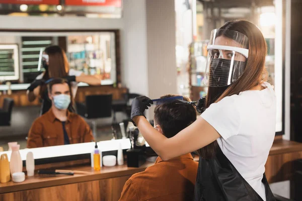 Hairdresser in face shield cutting hair of client in medical mask near blurred mirror reflection — Stock Photo