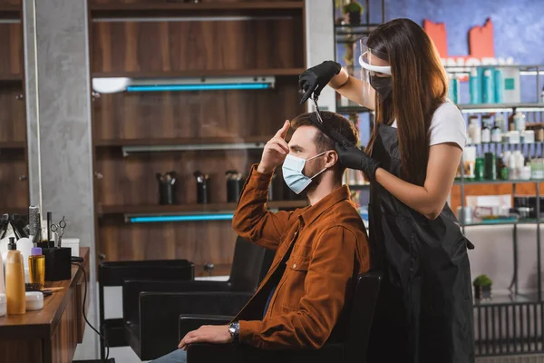 Seitenansicht eines Mannes, der mit den Fingern auf sein Haar zeigt, während der Friseur im Gesichtsschutz Haarschnitt macht — Stockfoto