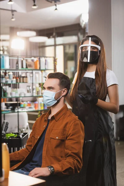 Peluquero en el escudo de la cara mirando a la cámara mientras está de pie cerca del cliente usando máscara médica - foto de stock