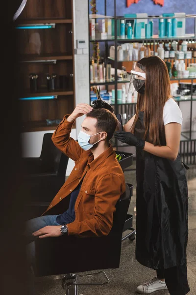 Man in medical mask touching his hair near hairdresser in face shield holding comb and scissors, blurred foreground — Stock Photo