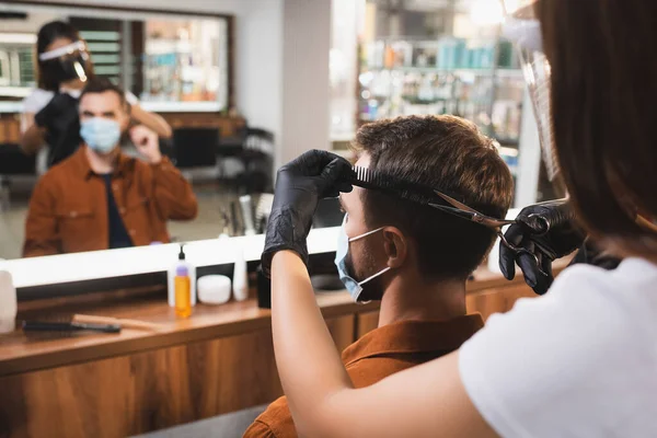 Hairdresser in face shield and latex gloves cutting hair of man with blurred mirror reflection — Stock Photo