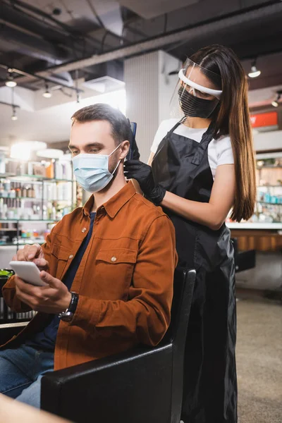 Homem em máscara médica usando smartphone enquanto barbeiro no escudo facial cortando seu cabelo — Fotografia de Stock