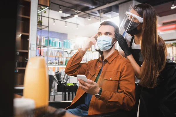 Uomo in maschera medica che tiene smartphone e punta con dito vicino parrucchiere in faccia scudo, banner — Foto stock