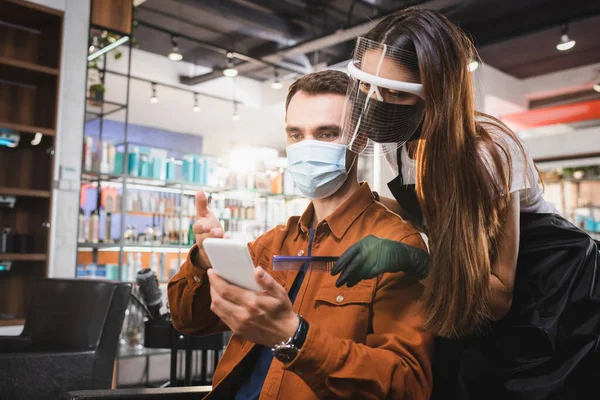 Barbiere in faccia scudo tenendo pettine vicino al cliente in maschera medica che punta con mano a smartphone — Foto stock