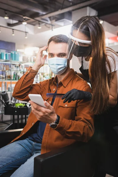 Peluquero en escudo facial apuntando con peine en el teléfono inteligente en las manos del hombre tocándose el pelo - foto de stock