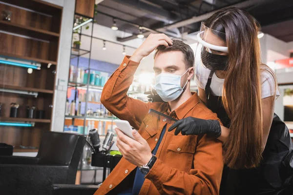 Barbeiro no rosto escudo e luvas de látex perto do cliente tocando seu cabelo e segurando smartphone — Fotografia de Stock