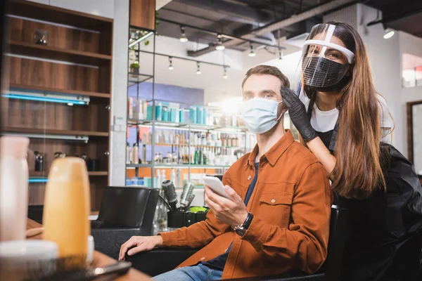Парикмахер в лице щит глядя на камеру рядом с человеком в медицинской маске держа смартфон, размытый передний план — стоковое фото