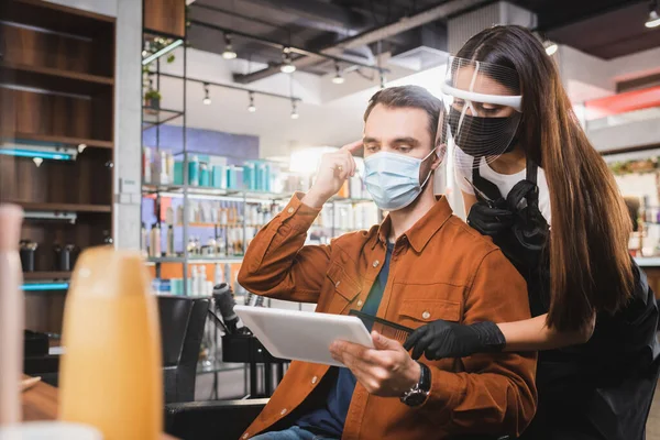 Man in medical mask holding tablet and pointing with finger at his hair near hairdresser holding comb — Stock Photo