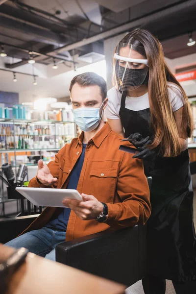 Uomo in maschera medica che punta con mano al tablet vicino parrucchiere in attrezzature protettive — Foto stock