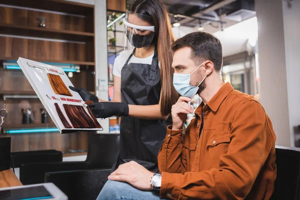 Coiffeur dans le bouclier facial et tablier montrant la palette de couleurs de cheveux à l'homme dans le masque médical — Photo de stock