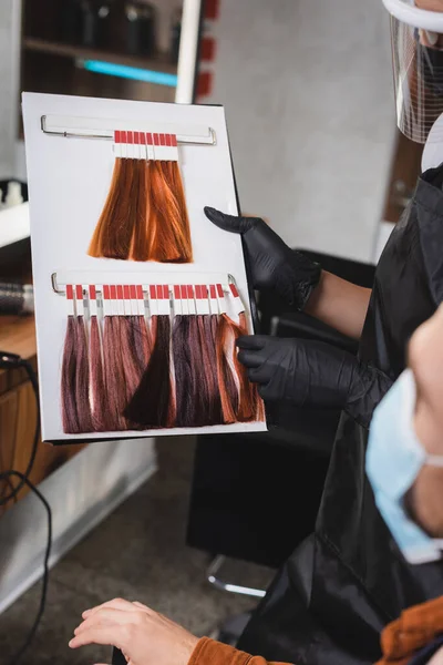 Cropped view of hairstylist in rubber gloves holding hair colors palette near client on blurred foreground — Stock Photo