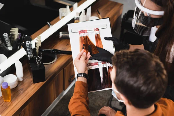 Hairdresser in face shield showing hair colors palette near client on blurred foreground — Stock Photo