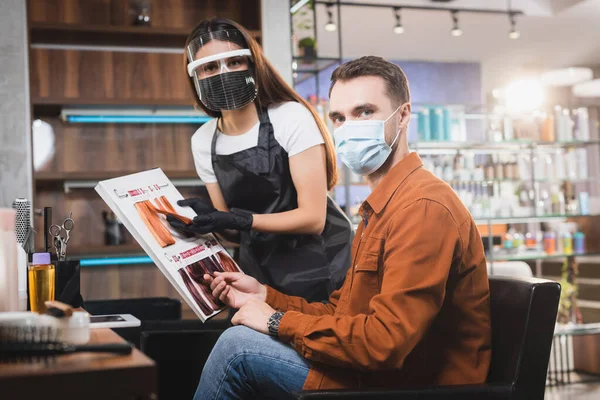 Cabeleireiro em equipamento de proteção olhando para a câmera enquanto segurando paleta de cores de cabelo perto do homem em máscara médica — Fotografia de Stock