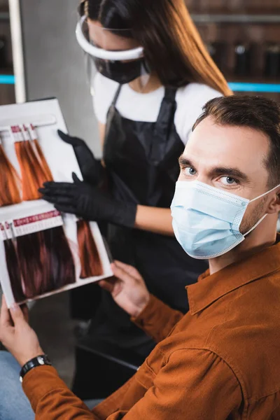 Homem em máscara médica olhando para a câmera enquanto segurando paleta de cores do cabelo com cabeleireiro no fundo borrado — Fotografia de Stock