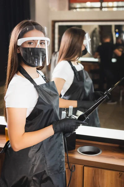 Young barber in face shield, apron and latex gloves holding curling iron on blurred background — Stock Photo