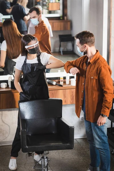 Junger Friseur mit Gesichtsschutz und Schürze stößt mit Mann in medizinischer Maske gegen Ellbogen — Stockfoto