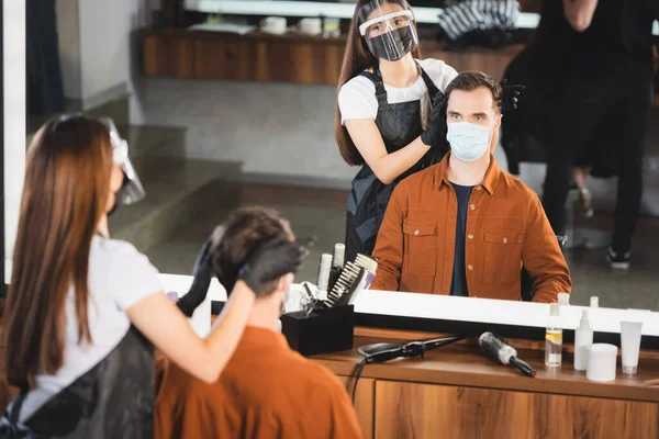 Mirror reflection of barber in face shield and latex gloves near client in medical mask on blurred foreground — Stock Photo