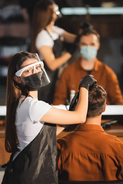 Парикмахер в лице щит глядя на камеру при прикосновении волос клиента на размытом фоне — стоковое фото