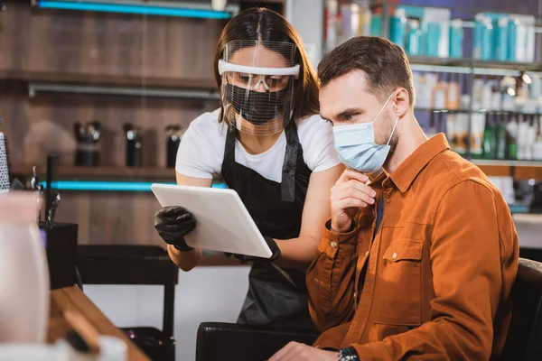 Peluquería en el escudo facial y guantes de látex que sostiene la tableta digital cerca del cliente en la máscara médica, borrosa primer plano - foto de stock