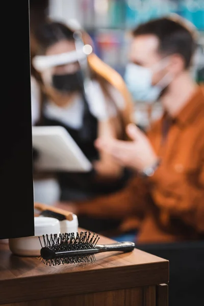 Comb and container of cosmetics on desk near barber and client in personal protective equipment on blurred background — Stock Photo