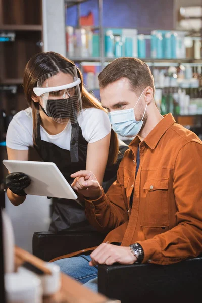 Barber in face shield and latex gloves holding digital tablet near client pointing with finger, blurred foreground — Stock Photo