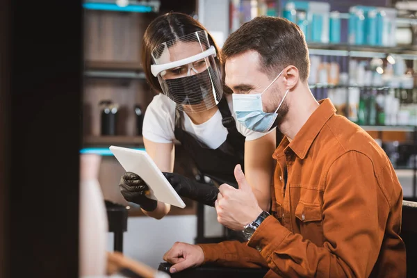 Friseur im Gesicht Schild hält digitales Tablet an Client zeigt Daumen nach oben, verschwommener Vordergrund — Stockfoto